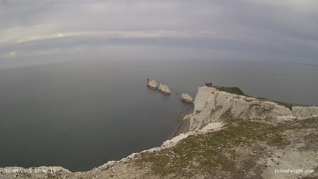 A coastal scene featuring high white cliffs at the forefront, with a rocky shoreline extending into the water below. In the distance, there are three prominent white rock formations emerging from the sea, one of which has a red and white lighthouse structure on it. The sky is overcast with gray clouds, and the water appears calm with a slight ripple, reflecting the muted tones of the sky. A few boats can be seen in the distance. The image captures a serene maritime landscape.