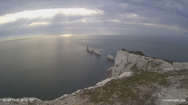 A scenic view of a coastline with white chalk cliffs meeting a calm blue sea. In the distance, a pair of rocky formations can be seen, with one featuring a red and white striped lighthouse. The sky is partially cloudy with rays of sunlight breaking through, illuminating the water. A grassy area is in the foreground, leading up to the cliffs. The atmosphere is serene and peaceful.