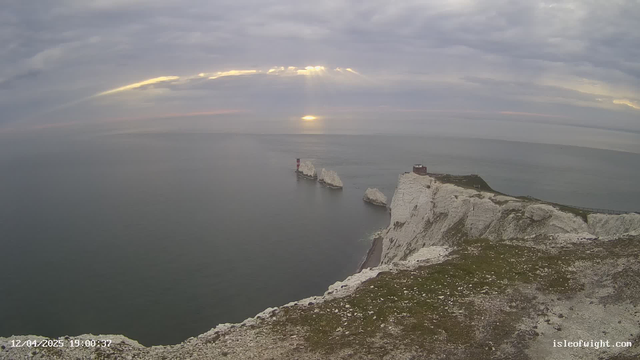 A panoramic view of a coastline featuring white chalk cliffs and a calm sea. In the foreground, there is rocky terrain with patches of green grass. To the right, a small building sits at the edge of the cliff, and farther out in the water, two prominent rocky outcrops are visible, one with a red and white striped lighthouse. The sky is overcast with gray clouds, and beams of sunlight break through, reflecting on the water's surface.