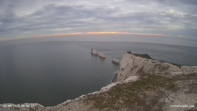 A coastal scene featuring steep white chalk cliffs bordering a calm sea. In the background, several rocky outcrops emerge from the water, with one marked by a red lighthouse. The sky is overcast with grey clouds and a faint hint of sunset colors illuminating the horizon. The foreground includes some green grass on the cliff edge, creating a natural setting above the water.