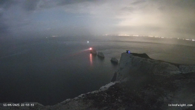 A nighttime coastal scene with rocky cliffs extending over dark water. In the distance, faint city lights are visible across the water, and a small boat emits a red light, creating a reflection on the water's surface. The sky is overcast with clouds, giving the scene a misty atmosphere. A structure is positioned on the cliff, possibly a lookout point, with dim light visible. The timestamp at the bottom indicates the date and time of the image.