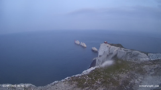 A panoramic view of the ocean under a gray sky, with rocky cliffs on the right side. Several natural rock formations are visible in the water, and a small red structure is seen atop one of the cliffs. The scene appears calm and serene, with gentle waves lapping at the shore. The landscape is mostly in cool tones, creating a tranquil atmosphere.
