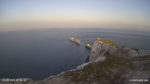 A coastal view during dawn shows a rocky shoreline with white cliffs on the right. Several large, white rock formations protrude from the calm blue sea, with a small red lighthouse visible on one of the rocks. The sky above is a gradient of light blue to pale yellow, indicating the early morning light. In the distance, a small sailing boat can be seen on the water. The foreground features grassy terrain, hinting at a natural environment, while the overall scene conveys tranquility.