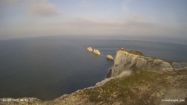 A coastal scene featuring cliffs resembling white chalk. In the background, there is a vast body of calm blue water under a light blue sky with some clouds. To the right, a small building is perched on top of the cliffs, and further out in the water, there are two larger rocky formations. A distant boat is visible on the water's surface. The foreground is grassy with rocky textures.