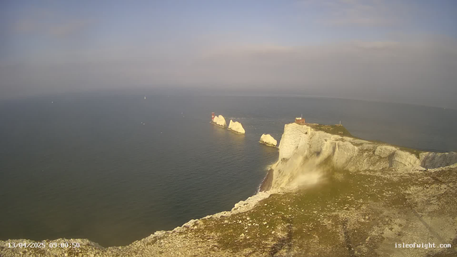 A panoramic view of the coastline featuring white chalk cliffs and a calm sea. In the foreground, rocky terrain with sparse vegetation leads to the cliff's edge. Three structures with red roofs are visible on the water's edge, set against a backdrop of the sea and a clear, partly cloudy sky. The water has small ripples, and a few boats are seen in the distance. The image captures a serene natural landscape.