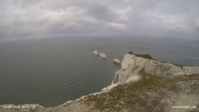 A panoramic view of a rocky coastline under a cloudy sky. The image features white chalk cliffs on the right, with a small building atop one of the cliffs. In the distance, several rock formations rise from the sea, one of which has a red and white lighthouse. The water is calm with a few small boats visible, and the horizon line blends into the overcast sky. The scene conveys a sense of natural beauty and tranquility.