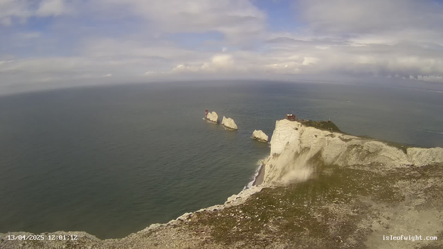 A coastal scene featuring rugged white cliffs rising from a deep blue ocean under a partly cloudy sky. In the distance, three rock formations are visible, with structures resembling lighthouses on top. The foreground shows grassy terrain with scattered rocks, leading to the edge of the cliffs. The sky is bright with clouds, creating a serene and picturesque maritime landscape.