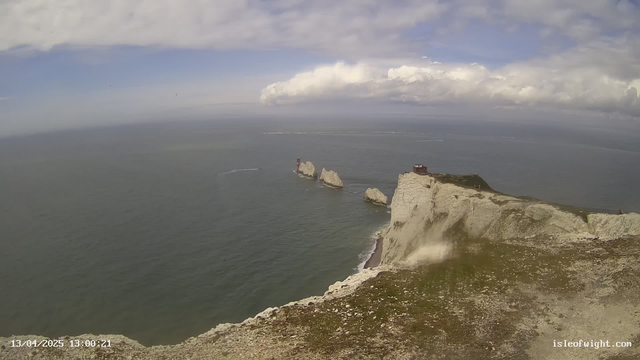 A coastal view featuring white chalk cliffs that drop into the blue sea. In the distance, several rocky outcrops emerge from the water, with a small red building on one of them. The sky is partly cloudy, with patches of blue and soft white clouds. The foreground shows green grass and rocky terrain along the cliff's edge.