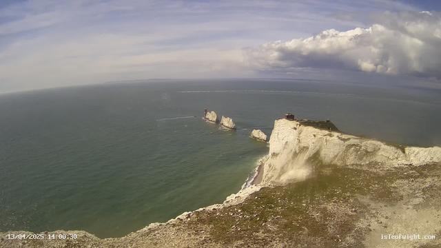 A coastal scene featuring rugged cliffs and a rocky shoreline. The water is calm and appears blue, stretching out towards the horizon. In the background, a few cloud formations are visible in a predominantly clear sky. A building is situated on top of the cliff, and two rock formations can be seen jutting out into the water. The foreground includes a grassy area with scattered stones.