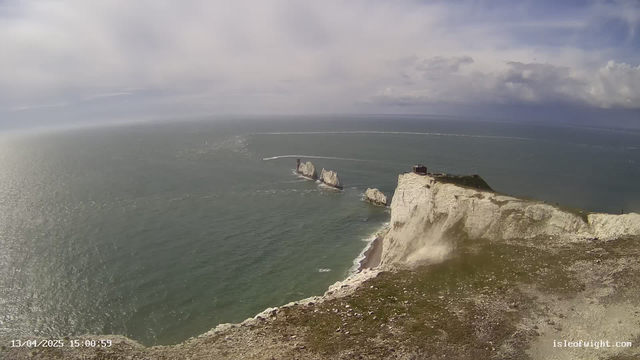 A panoramic view of a coastline featuring white chalk cliffs that drop steeply into the ocean. In the distance, several rocky formations partially emerge from the water. The sea appears choppy with small waves, reflecting sunlight. A few clouds are scattered across the sky, and a small building is visible at the top of the cliff near the edge. The image captures a sense of natural beauty and rugged coastline.