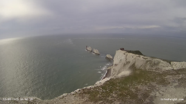 A wide view of a coastline featuring white cliffs submerged partly in the sea. The water appears calm with subtle waves, reflecting a muted light from overcast skies. Several rock formations rise from the water, some topped with structures resembling lighthouses. The foreground shows grass and rocky terrain leading to the cliff's edge. In the far distance, the horizon meets the cloudy sky. A time and date stamp is visible in the lower left corner, indicating the image was taken on April 13, 2025, at 16:00:18.