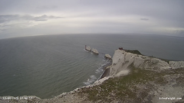 A coastal scene featuring white chalk cliffs rising sharply from the ocean. In the distance, two rocky formations are visible in the water, with a red and white lighthouse on one. The sky is overcast with gray clouds, and the water appears choppy, reflecting the light. The foreground shows grassy terrain, with some scattered rocks and a clear pathway leading toward the cliff edge.