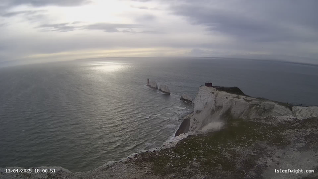 A view from a cliff overlooking a wide, calm ocean. The sea appears to be gently rippling, reflecting soft gray and silver light from a cloudy sky. In the distance, white chalk cliffs rise from the water, and one of the cliffs has a small, red lighthouse on it. The foreground shows a grassy, rocky edge leading down towards the ocean. The image has a tranquil and moody atmosphere, indicating it is late in the day.