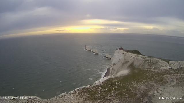 A coastal landscape view shows steep white cliffs descending into a choppy sea. In the distance, several rock pillars emerge from the water, with a lighthouse positioned on one of them. The sky is overcast with a blend of gray and blue hues, and the sun is setting on the horizon, casting a soft glow. The foreground features green grass and small stones scattered along the cliff edge.