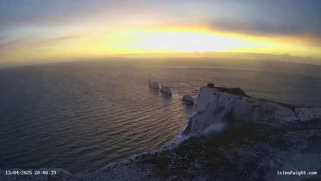 A scenic view of a coast at sunset. The ocean is calm, reflecting hues of orange and yellow from the sky. On the right, white chalk cliffs rise above the water, with a dark structure at the top. In the distance, several rock formations jut out from the water. The scene is tranquil, with a few clouds scattered in the sky.
