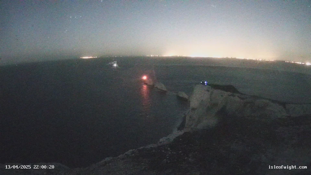 A twilight scene depicting a coastal view at night. The sea is calm, reflecting faint lights. In the distance, a coastal city can be seen with a line of lights along the horizon. To the right, there are rocky cliffs with a dim glow from a light source, possibly a beacon or a small vessel. The area is sparsely illuminated, creating a serene atmosphere. The date and time are displayed in the lower left corner.