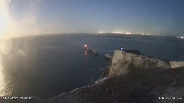 A webcam view of a coastal scene at dawn. The image shows a cliff on the right side, with a light shining from a structure at the edge. Below, there are rocky formations jutting into the water. The ocean appears dark and calm, reflecting some light. A faint city skyline is visible in the background, illuminated by distant lights. The date and time are displayed in the bottom left corner.