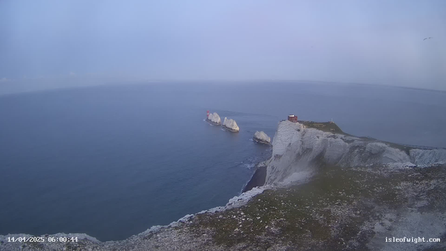 A panoramic view of a coastal area featuring steep white cliffs dropping into a calm sea. In the distance, there are several small rocky islands with red lighthouses. The sky is overcast, with a grayish-blue hue dominating the scene. Closer to the foreground, there is a green grassy area at the top of the cliff, with a pathway and some scattered rocks. The image captures a serene, foggy atmosphere.