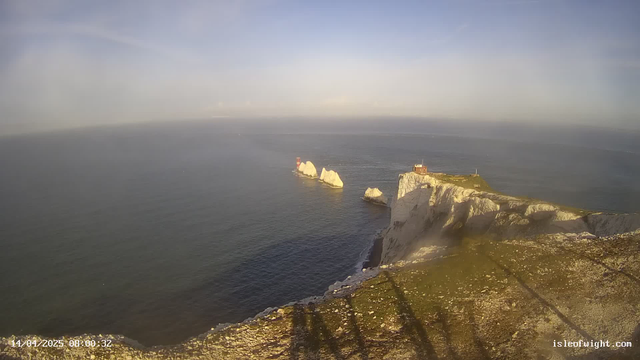 A coastal view featuring cliffs on the right side, extending into the ocean. Several large, white rock formations emerge from the water. A small red lighthouse is visible on one of the rocks, while another building, likely a lighthouse station, is located on the cliff's edge. The sky above is mostly clear with a hint of blue and soft clouds, while the ocean appears calm. Shadows cast by the cliff are visible on the ground.