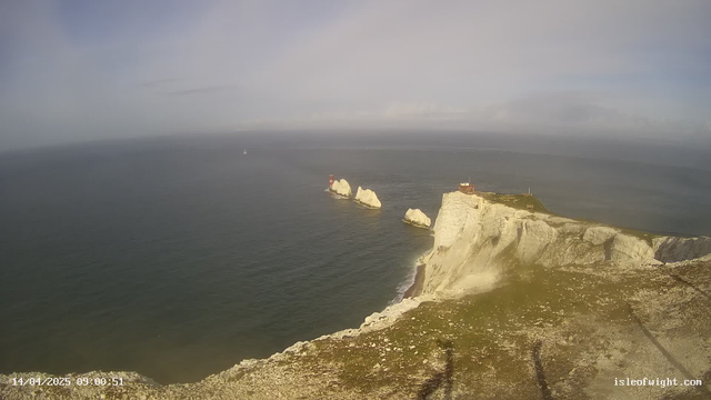 A coastal scene featuring white chalk cliffs that rise sharply from the ocean. In the distance, there are two rocky outcrops with red lighthouse structures on top. A small boat is visible on the water, and the sky above is partly cloudy, with a soft, light blue hue. The foreground includes rocky terrain and a grassy area.