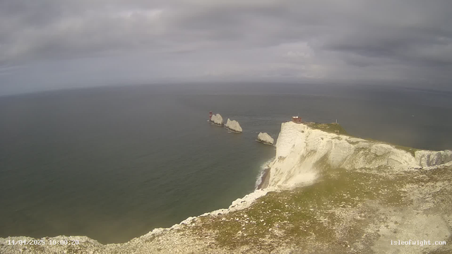 A coastal scene shows white chalk cliffs rising sharply above the sea, with dark waters stretching out to the horizon under a cloudy sky. There are two prominent red lighthouse structures positioned on rock formations offshore. The cliffs are dotted with patches of grass and small stones in the foreground. The overall atmosphere appears dramatic, highlighting the natural beauty of the area.