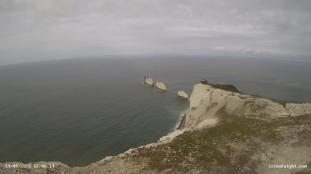 A coastal landscape featuring white chalk cliffs that overlook a calm sea. In the distance, there are three rocky outcrops with a structure on one of them. The sky is cloudy with gray tones, and the image captures a sense of tranquility. The foreground shows grassy terrain and scattered stones leading to the cliff edge. The scene has a serene atmosphere with no visible boats or wildlife.