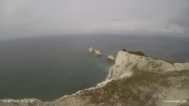 A coastal view featuring steep, white chalk cliffs on the right side, gradually sloping down to a rocky shoreline. In the distance, there are several smaller rock formations jutting out of the water. A calm sea reflects a cloudy sky, with patches of gray and blue. There are hints of green vegetation on the cliff tops and a few people walking along the paths. A structure resembling a building is visible near the top of the cliff. The date and time displayed at the bottom indicates the image was taken on April 14, 2025, at 13:01.