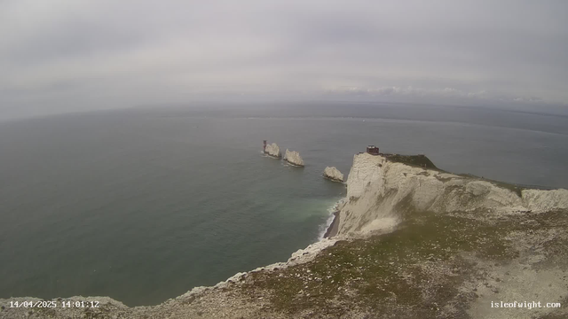A view from a cliff overlooking the ocean. The water appears calm and reflects the cloudy sky. In the distance, there are three prominent white rock formations protruding from the water, with a red lighthouse situated on one of them. The cliff edge is grassy and rocky, leading down to the shoreline. The scene conveys a sense of tranquility and natural beauty, with a predominantly gray color palette.