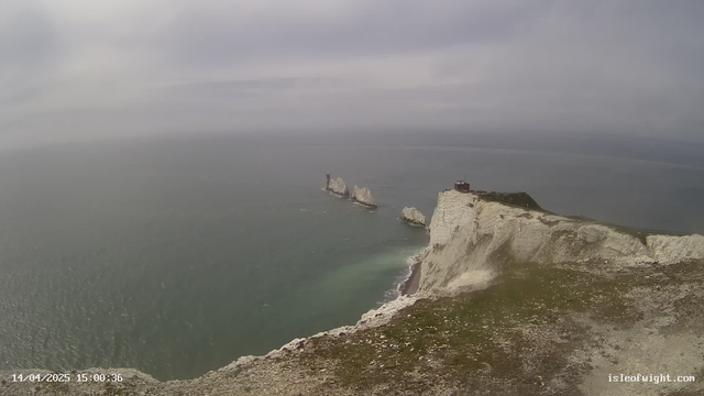 A scenic view of white chalk cliffs rising sharply from a calm sea, with several rocky outcrops visible in the water. The sky is overcast with gray clouds, and there is a small structure atop the cliff to the right. The foreground features grassy terrain leading down to the edge of the cliffs.