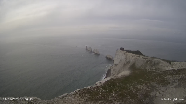 A coastal scene featuring white cliffs of chalk rising above the ocean. In the distance, three rocky outcrops are partially submerged in the water, with a small structure visible on the farthest rock. The sky is overcast, creating a grayish atmosphere. The shoreline curves along the bottom of the image, with some grass and rocks in the foreground. The ocean appears calm with gentle ripples.