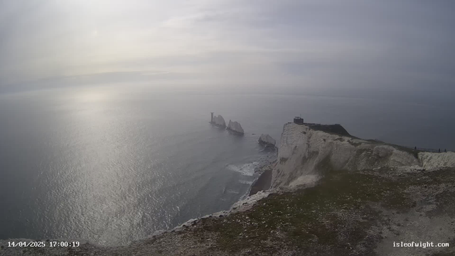 A coastal landscape featuring rugged white cliffs overlooking a calm sea. The water has a shimmering effect from the sunlight. In the distance, several large rock formations jut out from the water. A stone structure, possibly a lighthouse, is positioned on top of the cliffs, with a few people visible near the edge. The sky is mostly cloudy, creating a soft light over the scene.