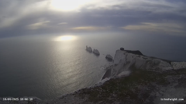 A coastal view featuring white chalk cliffs leading down to the sea. In the distance, several rugged rock formations emerge from the water. The sky is overcast with soft, gray clouds, and a faint light reflects off the calm surface of the ocean. The scene is serene, with a mixture of blue and gray tones dominating the landscape. A small building is situated at the edge of the cliffs, adding a sense of scale to the vast natural setting.