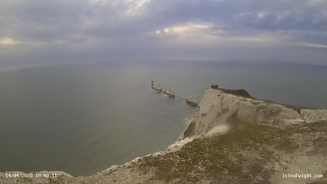A coastal view features rugged white cliffs descending to the sea. In the distance, a group of rocky islets is visible, with a red lighthouse situated on one of them. The sky is filled with clouds, creating a moody atmosphere. The water appears calm with gentle ripples, reflecting the overcast sky. There is a grassy area in the foreground, leading down towards the edge of the cliffs. The date and time are displayed in the lower left corner.