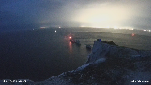 A dark, misty coastal scene at night. The foreground shows a rocky shoreline with a steep cliff on the right. In the water, several boats are visible, some with lights illuminating the surrounding fog. The background features a distant shoreline with faint lights from buildings or structures, creating a hazy glow in the atmosphere. The timestamp at the bottom indicates the image was taken on April 14, 2025, at 21:00:37.