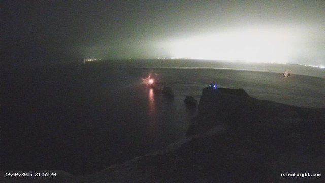 A coastal scene at night with low visibility. The foreground features dark rocky formations partially illuminated by a faint light. To the left, a small bright red light is visible, presumably from a boat or a buoy. In the background, faint lights from a distant shore are seen, creating a hazy glow against the dark sky. The overall atmosphere is misty and tranquil.