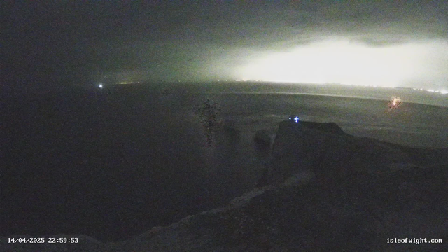 A night scene showing a rocky coastline illuminated by faint lights in the distance. The ocean appears dark and calm, with a prominent cliff edge on the right side. A small light, likely from a boat or equipment, is visible in the water. The sky is overcast, adding to the dim atmosphere of the scene. The timestamp at the bottom indicates the date and time of the image.