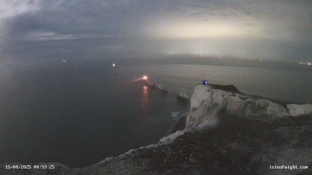 A foggy coastal scene at dawn, showing steep white cliffs leading down to the gently rippling sea. A small orange light reflects off the water near the edge of the cliffs, likely from a lighthouse, while faint lights from distant boats are visible in the mist. The sky is overcast with gray clouds, and the atmosphere appears calm and serene. The date and time display at the bottom indicates it’s April 15, 2025, at 4:59 AM.