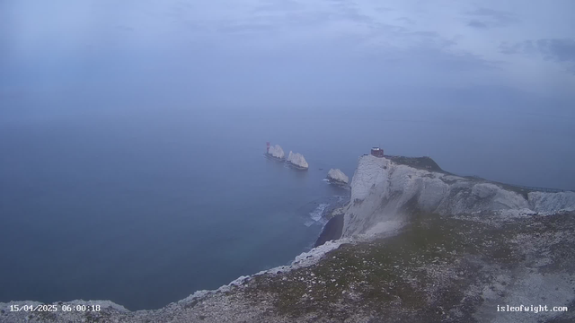 A coastal landscape featuring white chalk cliffs against a gray sky. In the distance, two large rock formations rise from the ocean, with a red lighthouse on one of them. The water appears calm and reflects the muted colors of the overcast sky. The foreground shows a grassy area atop the cliffs, leading to a steep drop into the sea. The scene is serene and slightly foggy, suggesting early morning light.