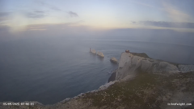 A coastal view shows steep white cliffs rising from the sea, with a lighthouse structure near the top of the cliffs. Below, several large rock formations jut out of the water, while the calm sea reflects soft hues of blue and gray. The sky above is slightly cloudy, with light peeking through, suggesting early morning light. The image captures a peaceful, natural landscape with a sense of tranquility.