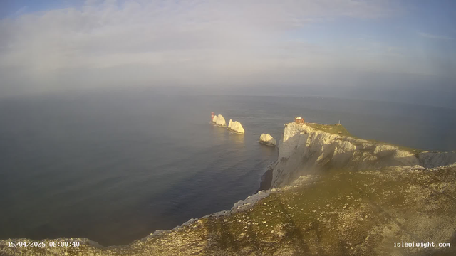 A coastal scene showing white chalk cliffs along the shoreline, with several rocky outcrops emerging from the water. A lighthouse with a reddish tower is positioned on one of the rocks, while another small structure with a red roof sits above the cliffs. The sea appears calm, reflecting the light of a cloudy sky, and the landscape includes some greenery at the cliff's edge. The image is timestamped in the lower left corner.