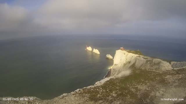 A scenic coastline view features white cliffs under a cloudy sky. In the distance, two rocky outcrops are visible, each topped with a red lighthouse. The sea is calm, reflecting shades of blue and green. The grassy cliff edge is in the foreground, while the horizon stretches out in the background.