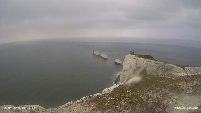A panoramic view of a coastal landscape featuring white cliffs overlooking a calm sea. In the distance, a series of rock formations emerge from the water, and a small lighthouse is visible atop one of the rocks. The sky is overcast with gray clouds, creating a moody atmosphere. The foreground shows grassy terrain leading to the cliff edge.