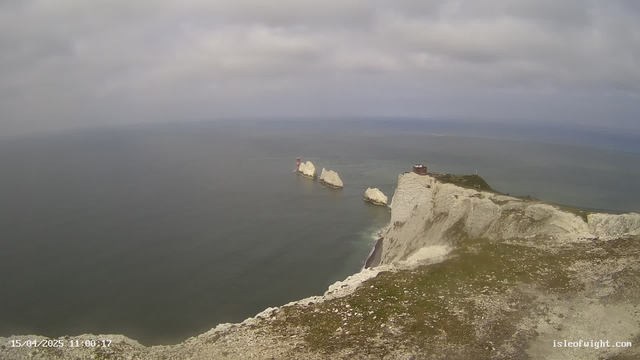 A scenic view of the coastline showing white chalk cliffs against a cloudy sky. In the distance, several rocky outcrops jut into the ocean, with one prominent structure displaying a red and white lighthouse. The ocean is calm, reflecting the gray sky, and some greenery is visible at the edge of the cliff in the foreground.