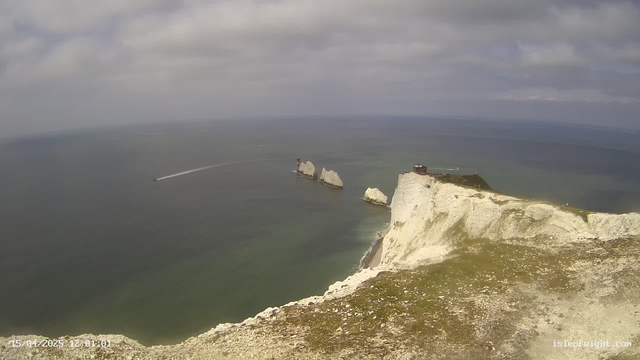 A scenic coastal view features steep white cliffs meeting a calm blue sea under a partly cloudy sky. In the distance, several small rocky formations with buildings on top are visible, along with a boat leaving a wake in the water. The shoreline includes a pebbly beach at the base of the cliffs, and there is a patch of green grass in the foreground. The image conveys a tranquil seaside atmosphere.