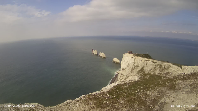 A scenic view of a coastal landscape featuring white chalk cliffs that drop into the calm blue sea. In the distance, there are several small rock formations protruding from the water. The sky is partly cloudy with patches of blue visible. The foreground includes greenery and rocky terrain near the cliff's edge, providing a natural, rugged appearance.