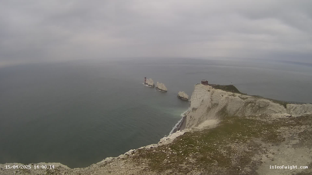 A cloudy sky above a calm sea with gentle waves. In the distance, three white rock formations rise from the water, the first one featuring a red and white striped lighthouse. The shoreline is rugged with steep white cliffs, and a small building is visible at the top of the cliff on the right. The foreground is covered with green grass and rocky terrain.