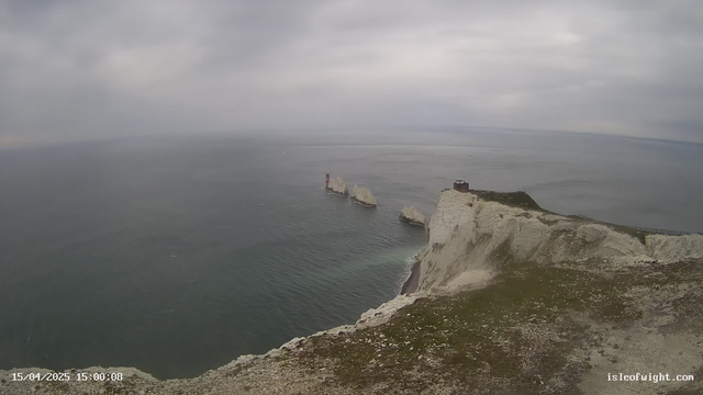 A coastal scene showing white chalk cliffs rising sharply from the sea. In the foreground, green grasses and rocky terrain lead up to the cliff edge. Below, waves gently lap against the base of the cliffs. Several rock formations jut out into the water, with a red lighthouse standing on one of the formations. The sky is overcast with a grayish hue, suggesting a cloudy day, and the ocean appears calm.