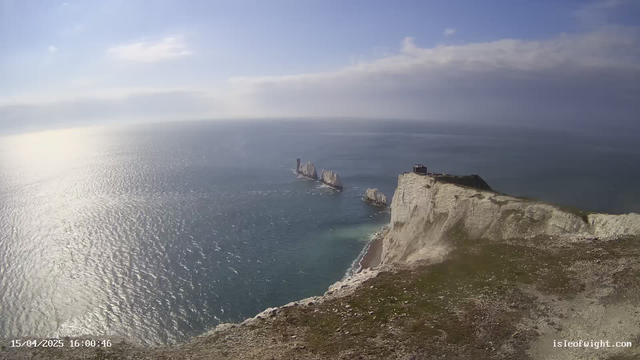 A scenic view of a coastline featuring steep white cliffs meeting the water. The ocean is calm and reflects sunlight, creating a shimmering effect across the surface. In the distance, several limestone formations rise from the sea. A small structure is visible at the cliff's edge, with grassy areas in the foreground. The sky is mostly clear with a few clouds scattered across the horizon.