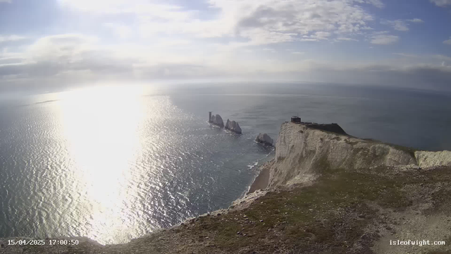 A panoramic view of a coastline featuring white chalk cliffs meeting the ocean. The sun is reflecting off the water, creating a shimmering effect. In the distance, jagged rock formations emerge from the water. The sky is partly cloudy, and there is greenery on the cliff's edge. A small structure is visible near the top of the cliff. The scene conveys a serene and natural landscape.