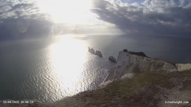 A panoramic view of a coastline featuring steep white cliffs descending into a shimmering sea. The water reflects sunlight, creating a glittering effect on the surface. In the distance, several rocky outcrops rise from the water, while a small, dark structure sits atop one of the cliffs. The sky is a mix of blue and gray clouds, conveying a moody atmosphere. The image captures the natural beauty of the area, including greenery at the cliff's edge.
